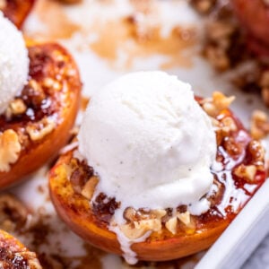 Baked peaches topped with brown sugar and pecans in a baking dish, topped with vanilla ice cream.