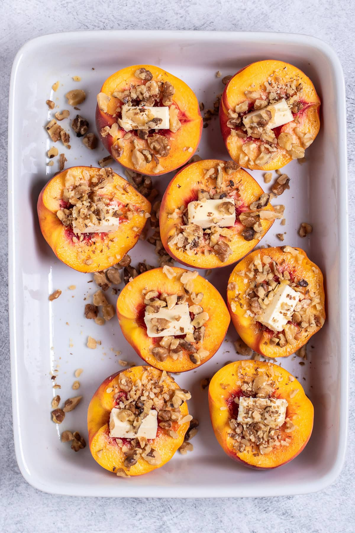 Peach halves open-side-up with butter and pecans in them in a baking dish.