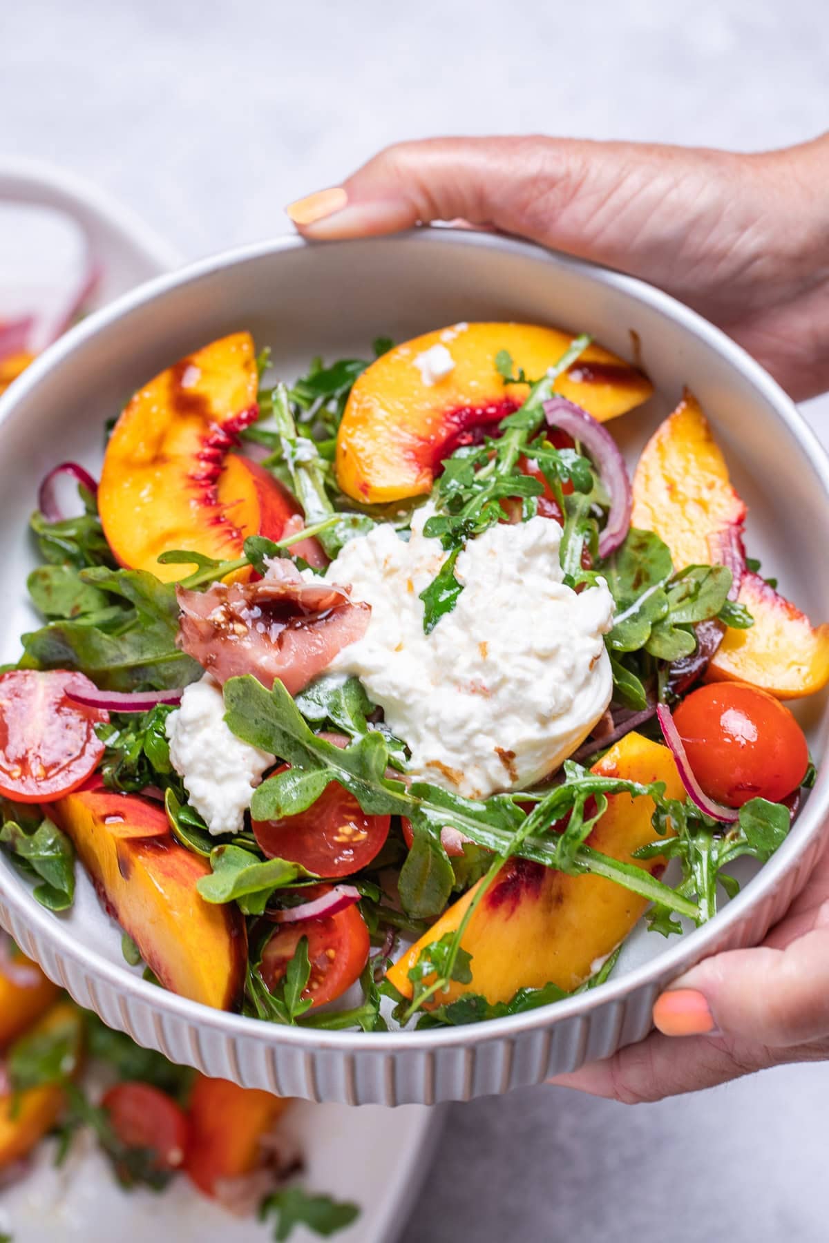 Two hands holding a bowl with peach burrata salad, with arugula, cherry tomatoes, red onion, peach slices, and burrata on top.