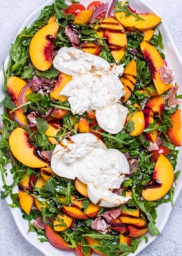 A serving platter on a table with peach burrata salad made with peaches, prosciutto, arugula, onion, and tomatoes tossed in dressing and topped with two big pieces of burrata, then drizzled with balsamic glaze.