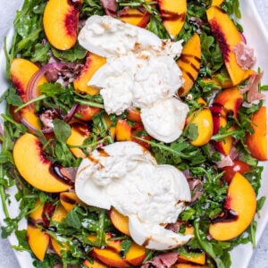 A serving platter on a table with peach burrata salad made with peaches, prosciutto, arugula, onion, and tomatoes tossed in dressing and topped with two big pieces of burrata, then drizzled with balsamic glaze.