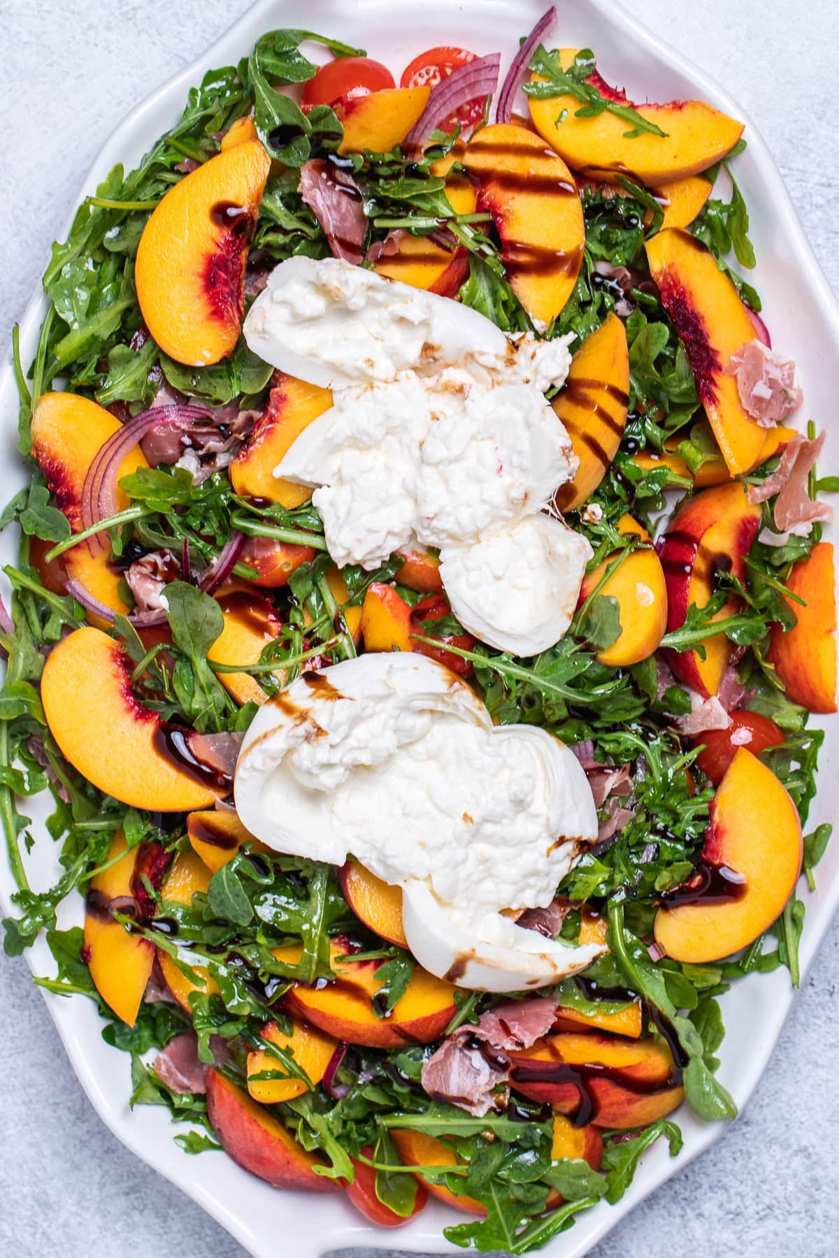 A serving platter on a table with peach burrata salad made with peaches, prosciutto, arugula, onion, and tomatoes tossed in dressing and topped with two big pieces of burrata, then drizzled with balsamic glaze.