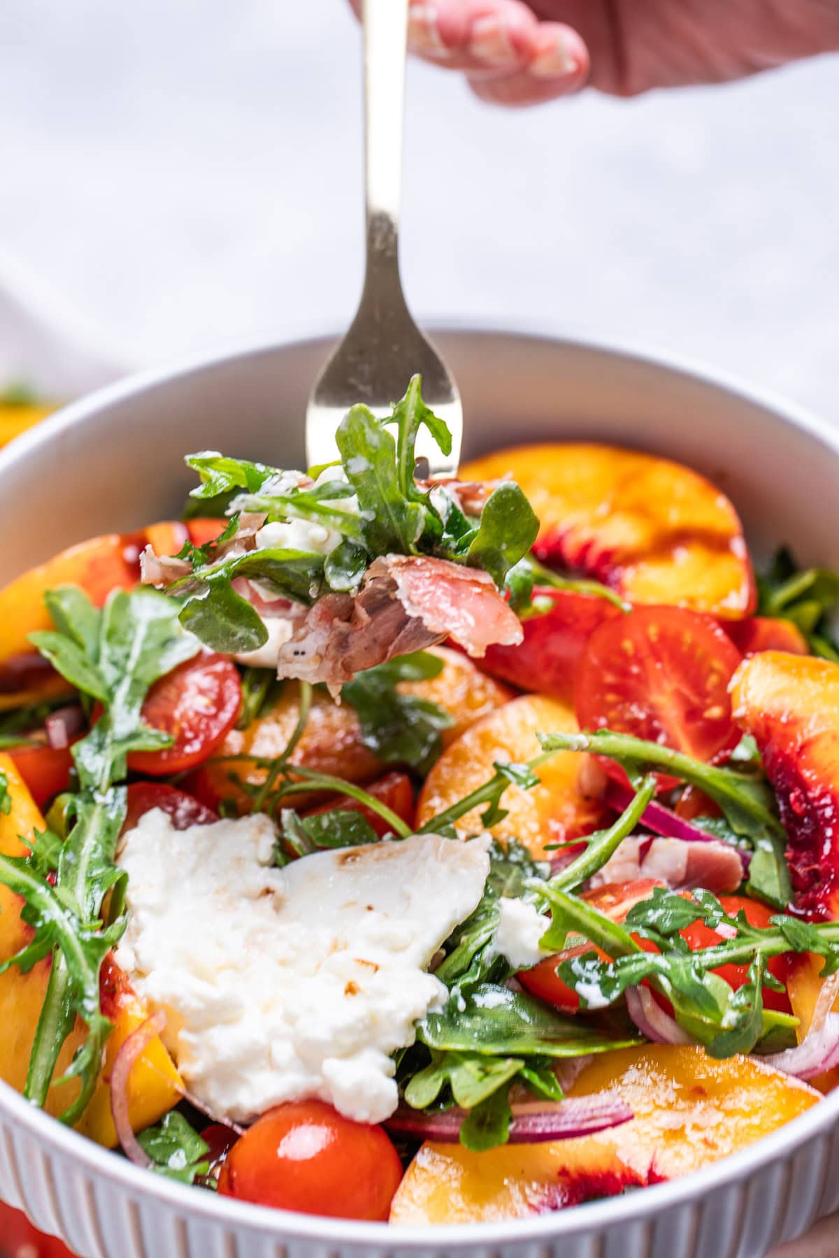 A bowl of peach burrata salad with arugula, cherry tomatoes, red onion, peach slices, and burrata on top, and a fork lifting some up.
