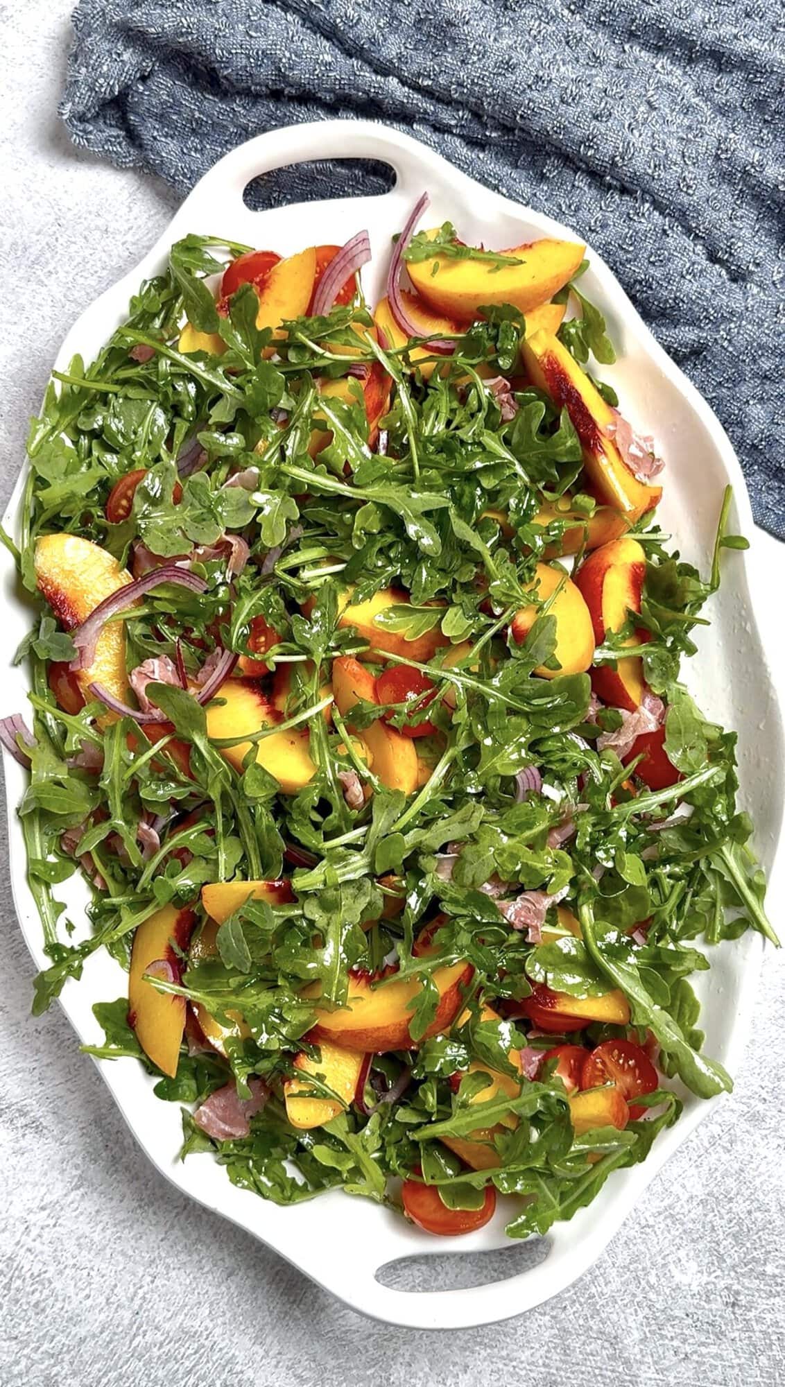 A serving platter on a table with peach burrata salad made with peaches, prosciutto, arugula, onion, and tomatoes tossed in dressing before adding other ingredients.