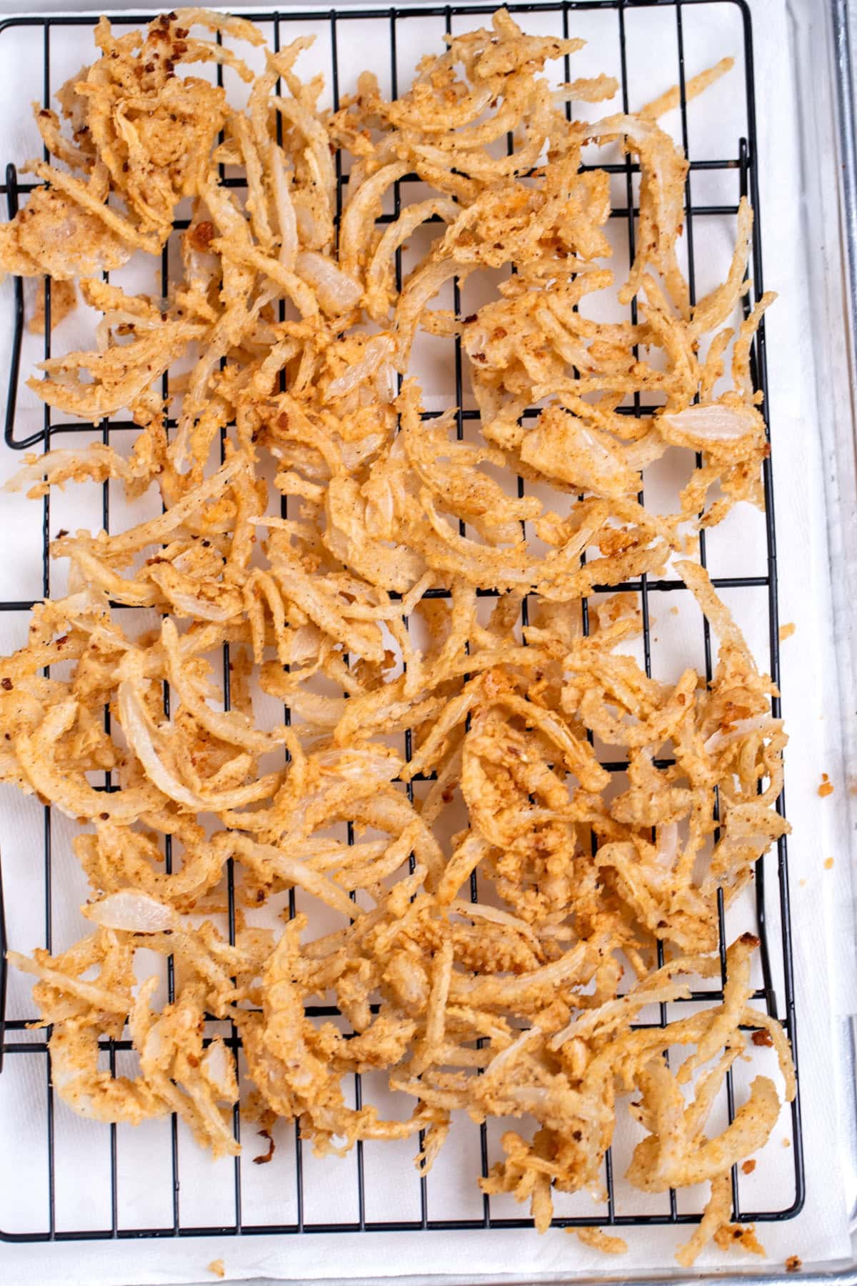A sheet pan lined with paper towels and a baking rack, with crispy fried onions cooling after being fried.