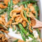 A wooden serving spoon lifting up some green bean casserole from a baking dish, with crispy fried onions on top.