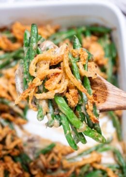 A wooden serving spoon lifting up some green bean casserole from a baking dish, with crispy fried onions on top.