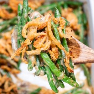 A wooden serving spoon lifting up some green bean casserole from a baking dish, with crispy fried onions on top.