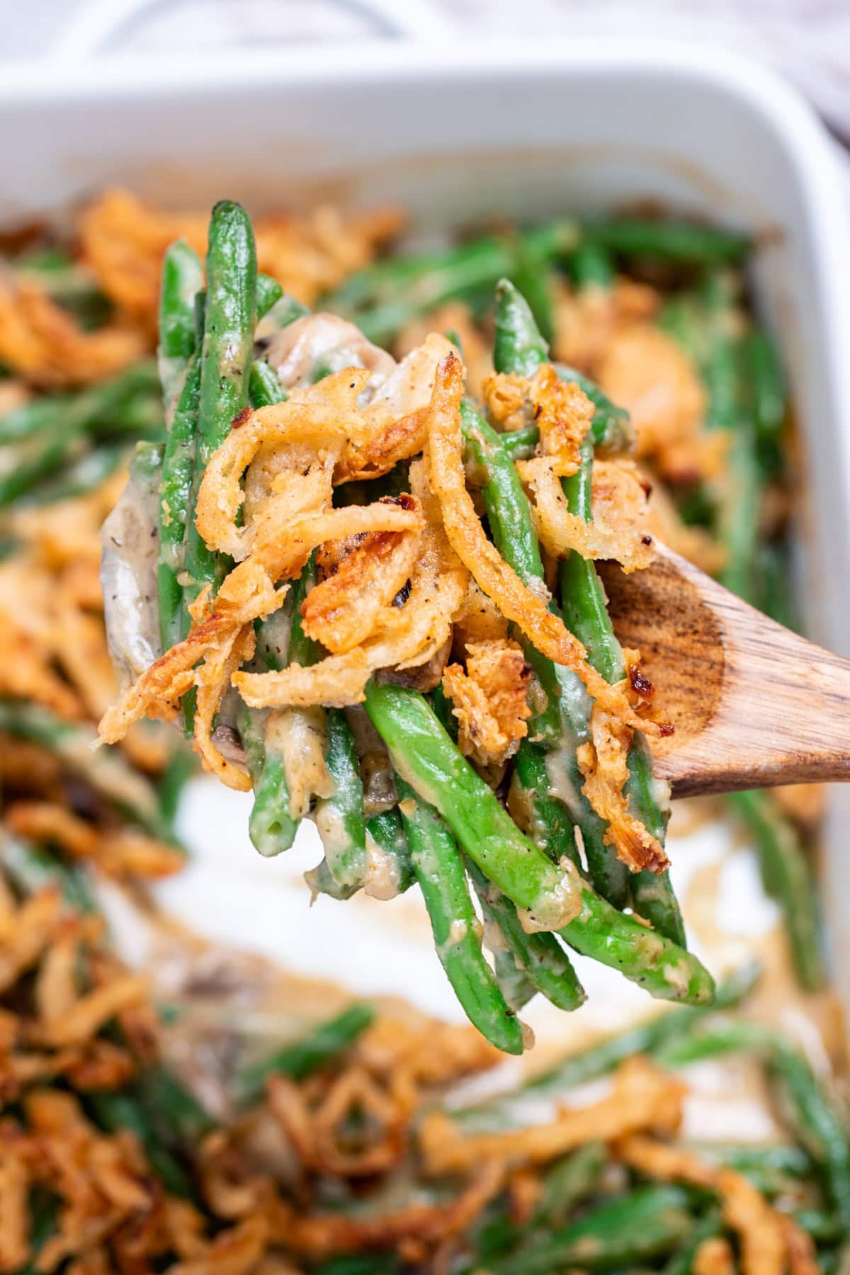 A wooden serving spoon lifting up some green bean casserole from a baking dish, with crispy fried onions on top.