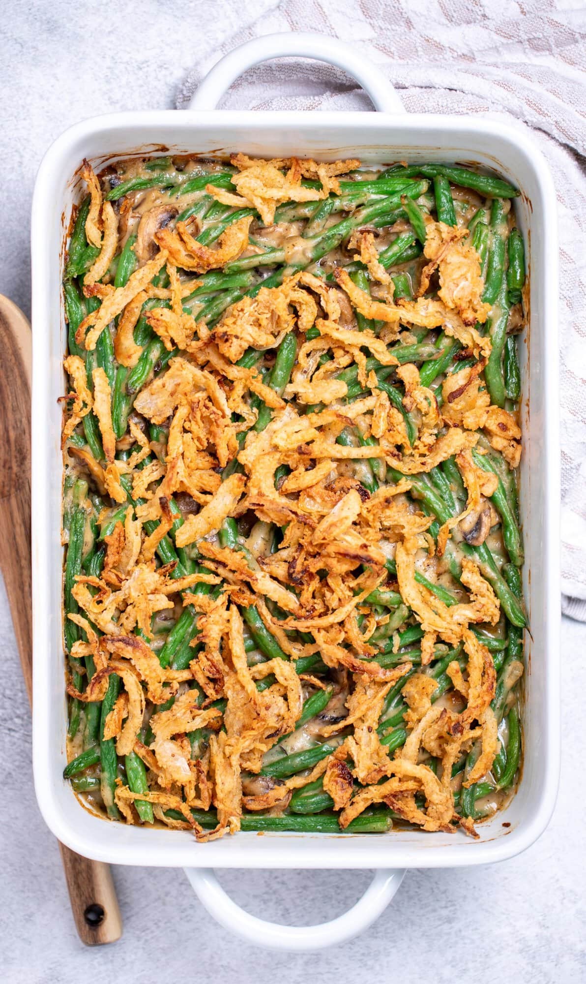 A casserole dish on a table next to a wooden spoon and a tea towel, full of gluten free green bean casserole topped with crispy fried onions.