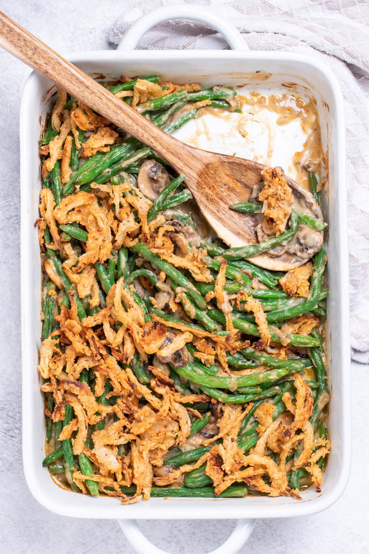 A casserole dish on a table next to a tea towel, full of gluten free green bean casserole topped with crispy fried onions, with a wooden serving spoon resting in an empty spot in the dish.