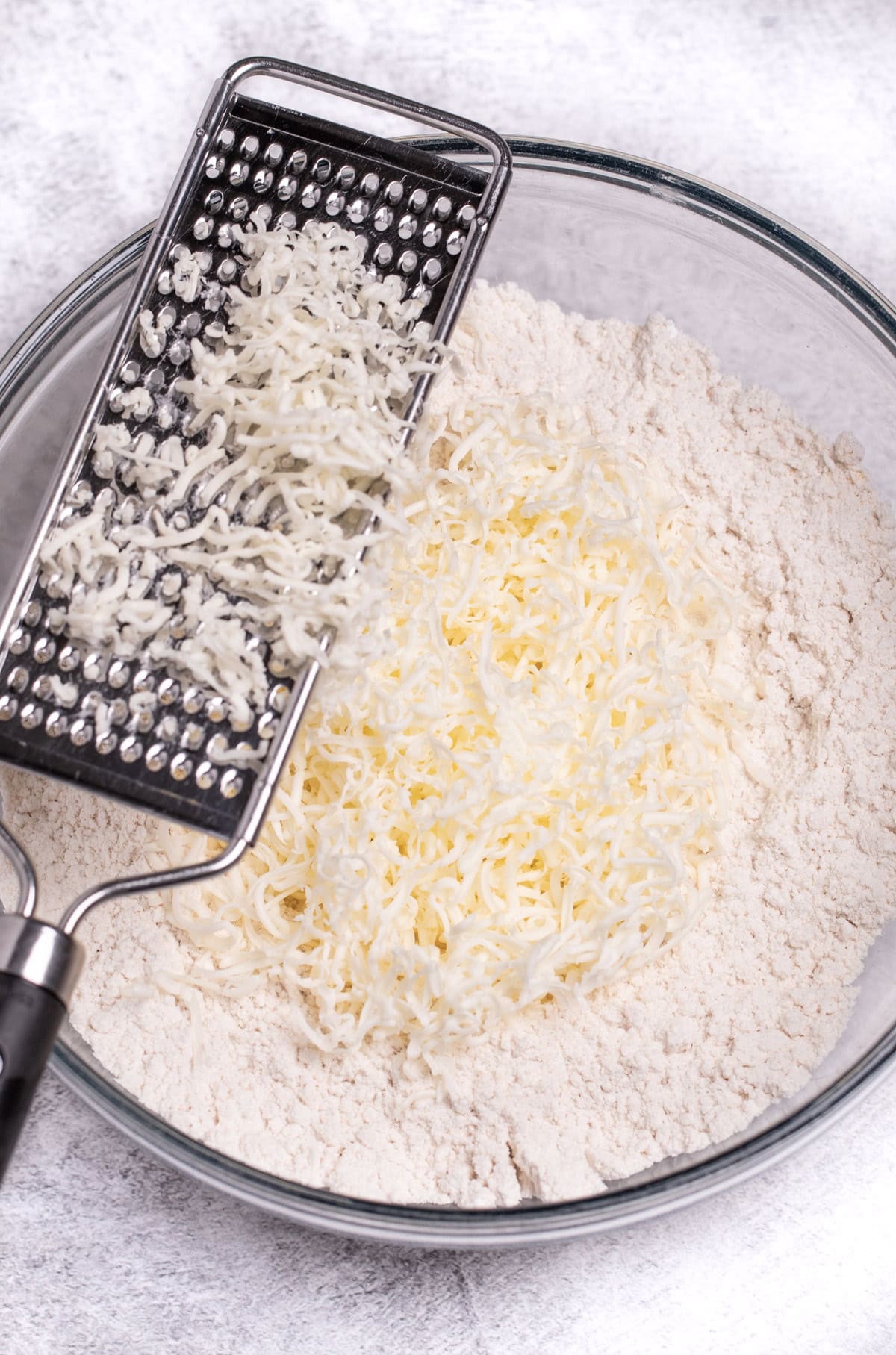 A mixing bowl of flour on a table with a grater resting on top, and butter that's been grated on top of the flour.