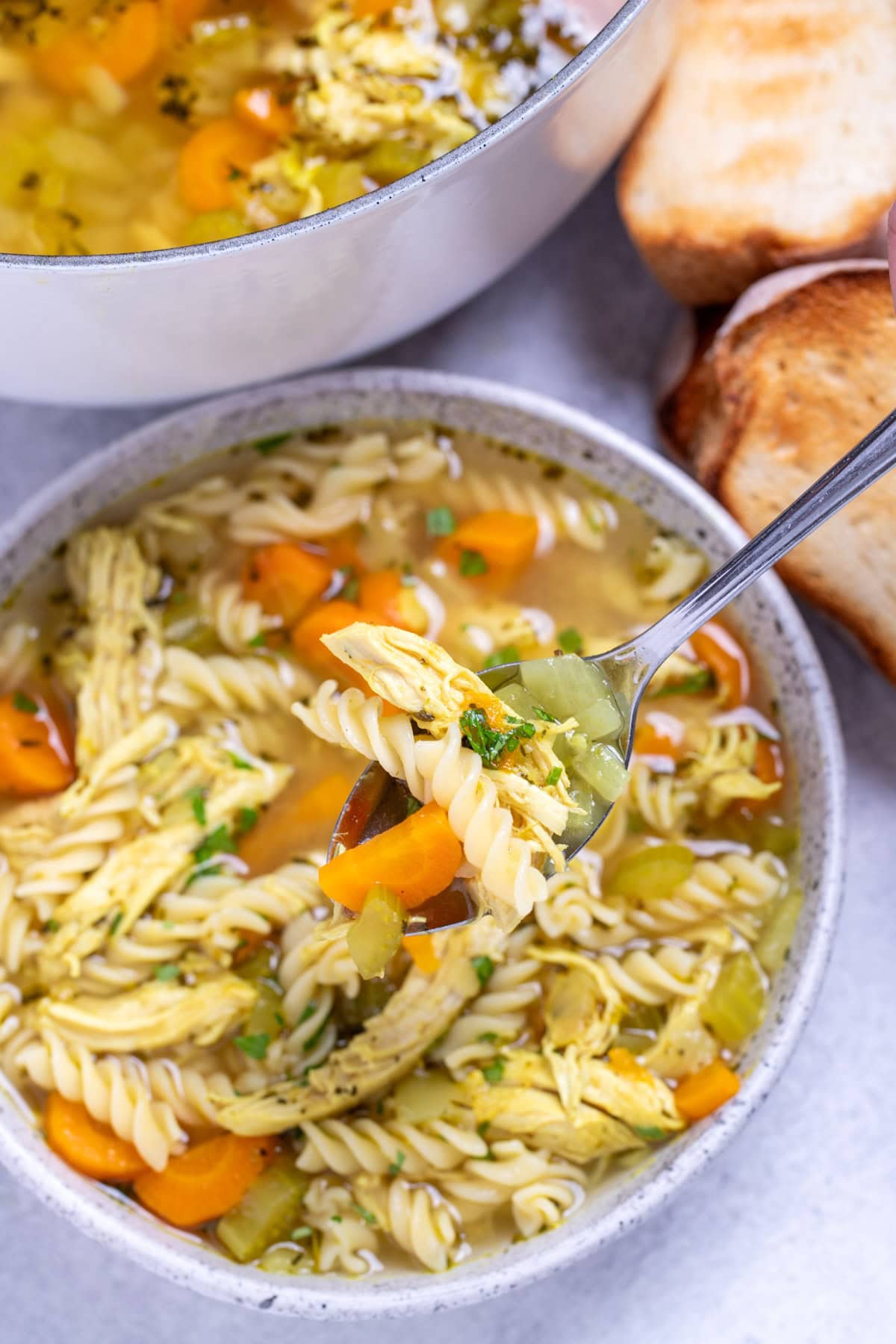 A bowl of gluten free chicken noodle soup in front of a dutch oven and toasted bread, with a spoon lifting some up.