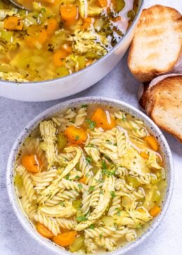 A bowl of gluten free chicken noodle soup in front of a dutch oven and toasted bread.
