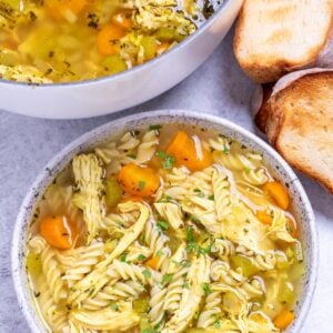 A bowl of gluten free chicken noodle soup in front of a dutch oven and toasted bread.