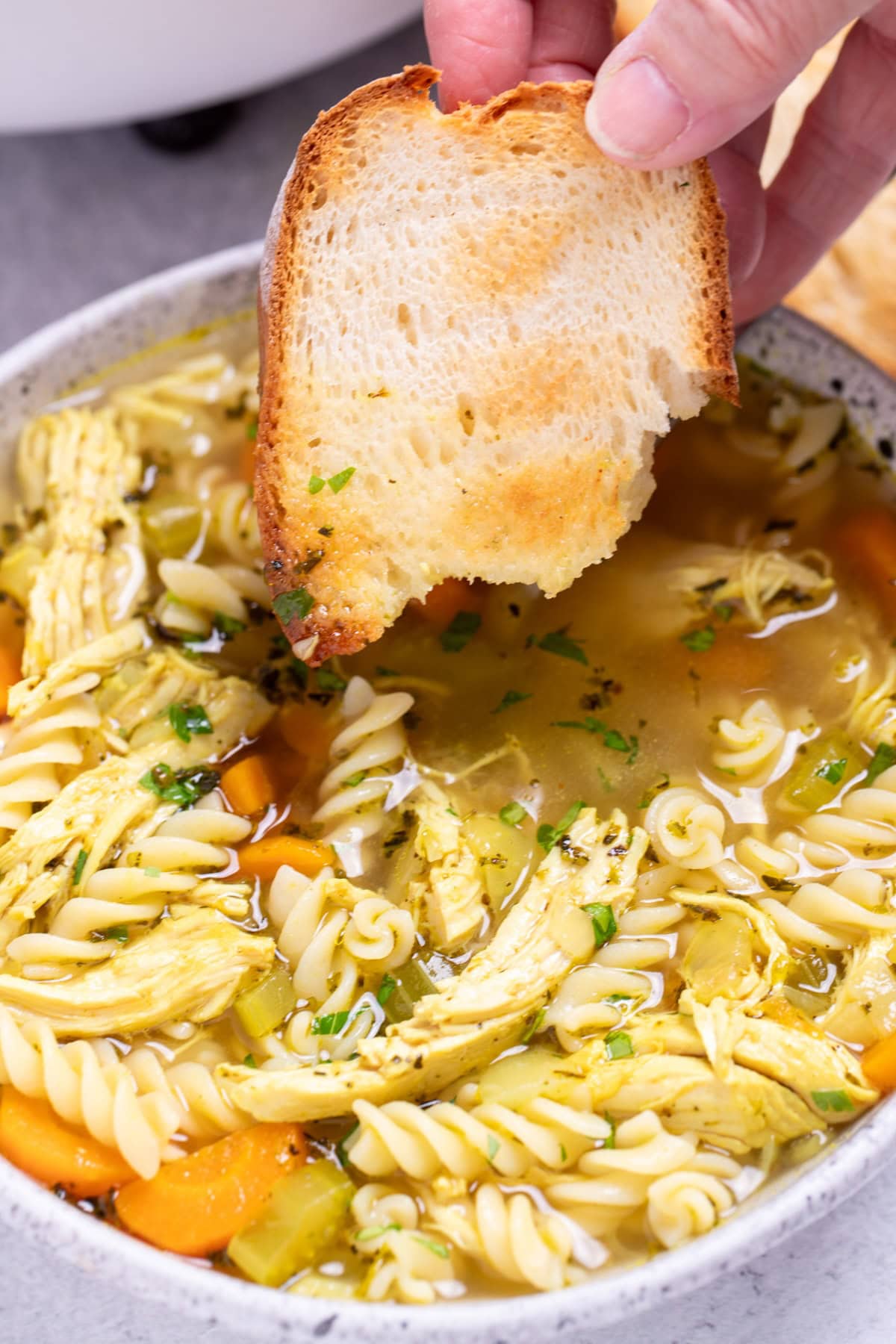 A bowl of gluten free chicken noodle soup with a hand dipping a piece of toasted bread into it.