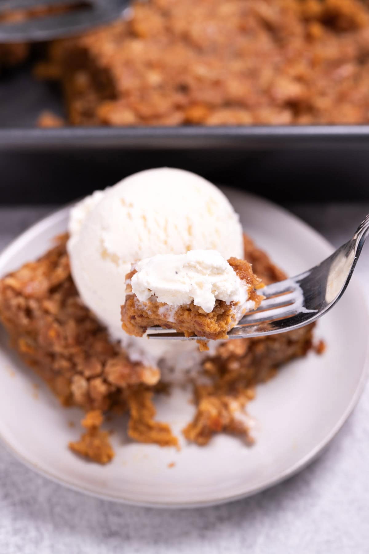 A plate with a serving of pumpkin crisp topped with ice cream in front of the baking dish of crisp, with a fork lifting some up.