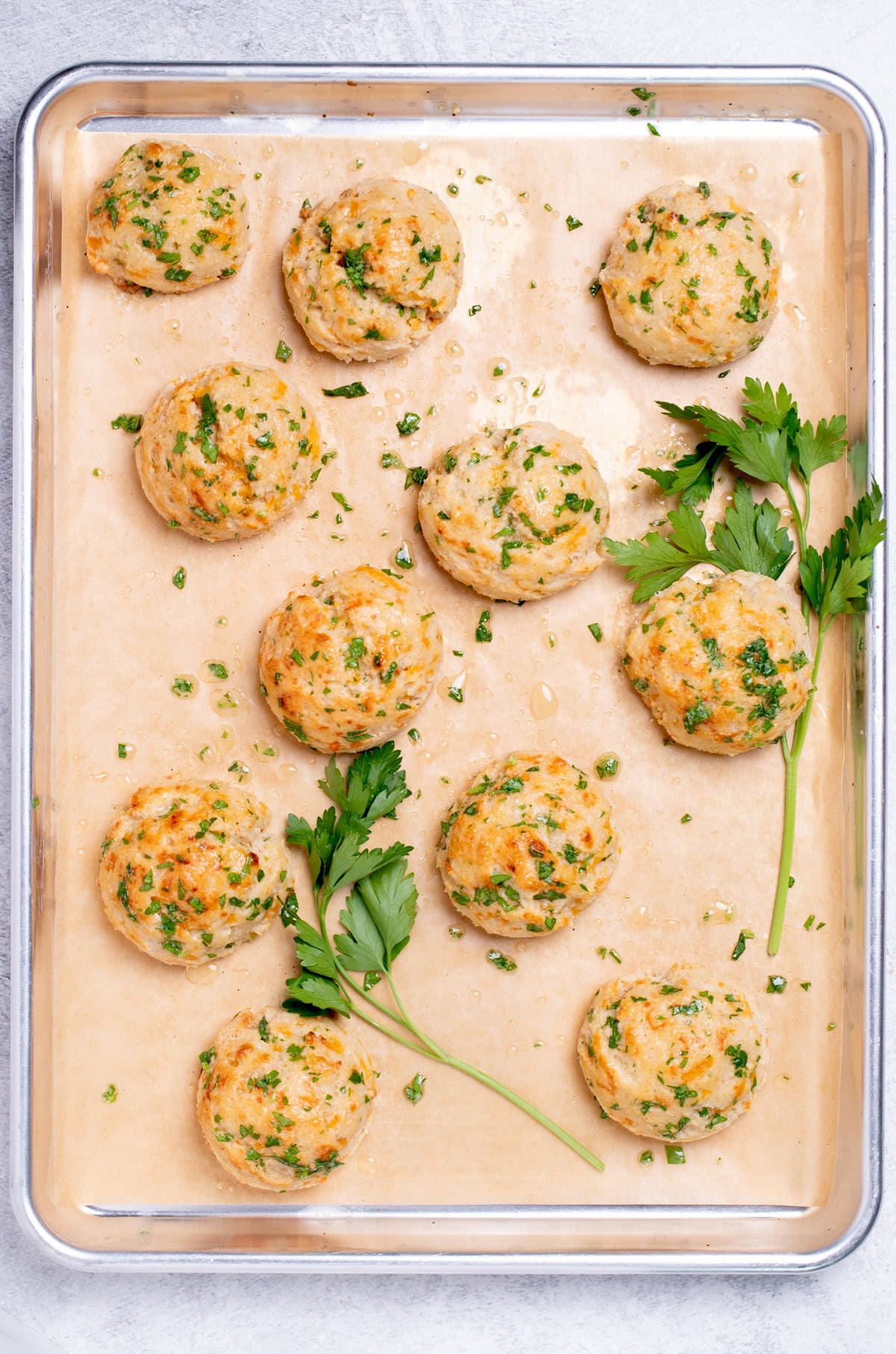 A sheet pan lined with parchment paper, with cheddar bay biscuits that have just been topped with seasoned herb butter, and pieces of parsley around them.