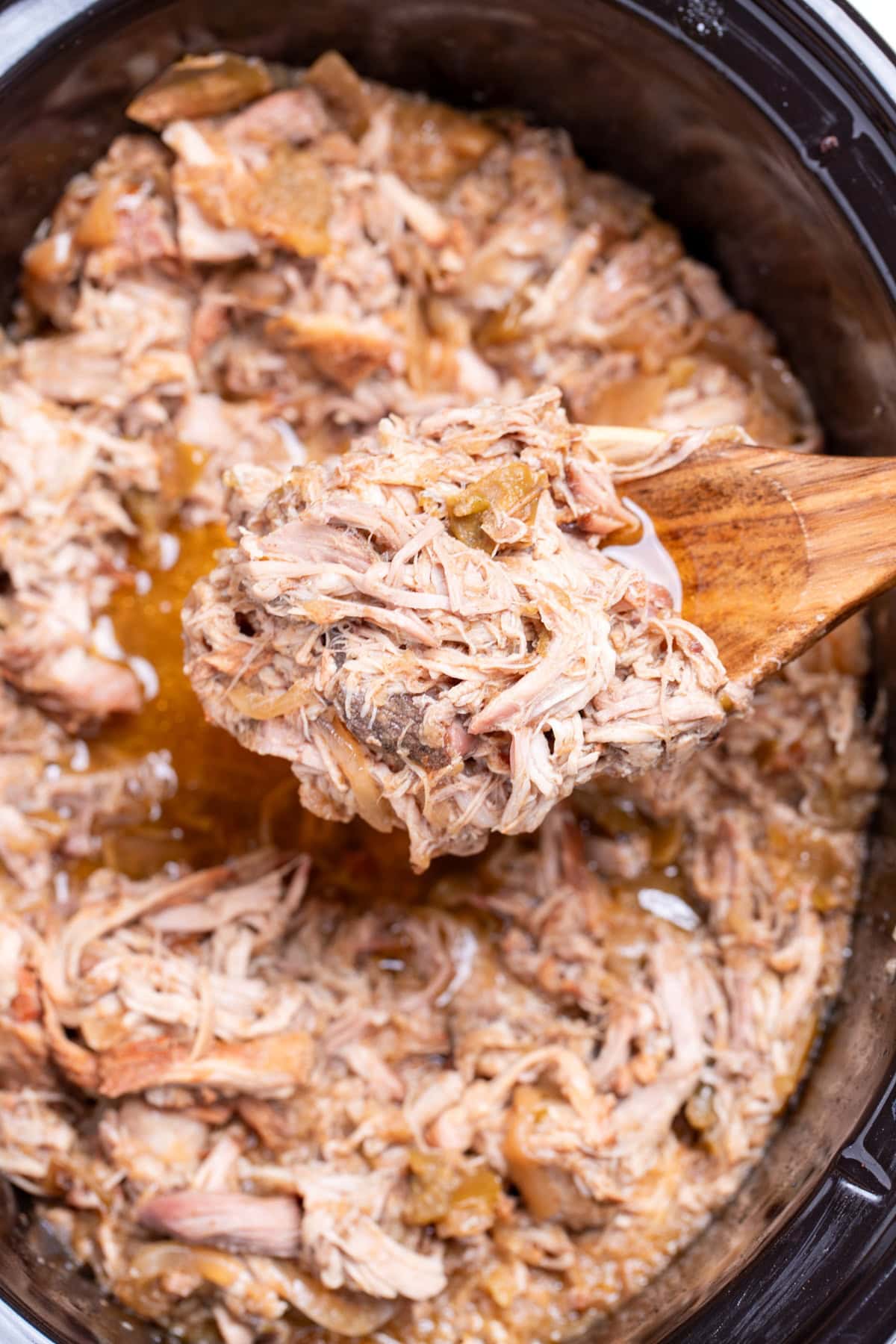 A wooden spoon lifting apple cider pulled pork out of a slow cooker.