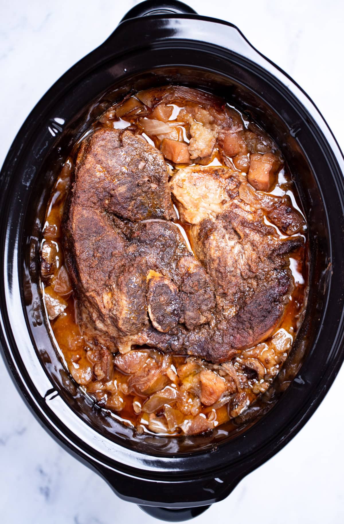 A slow cooker with a fully cooked pork shoulder in juices, apples and onions before being shredded.