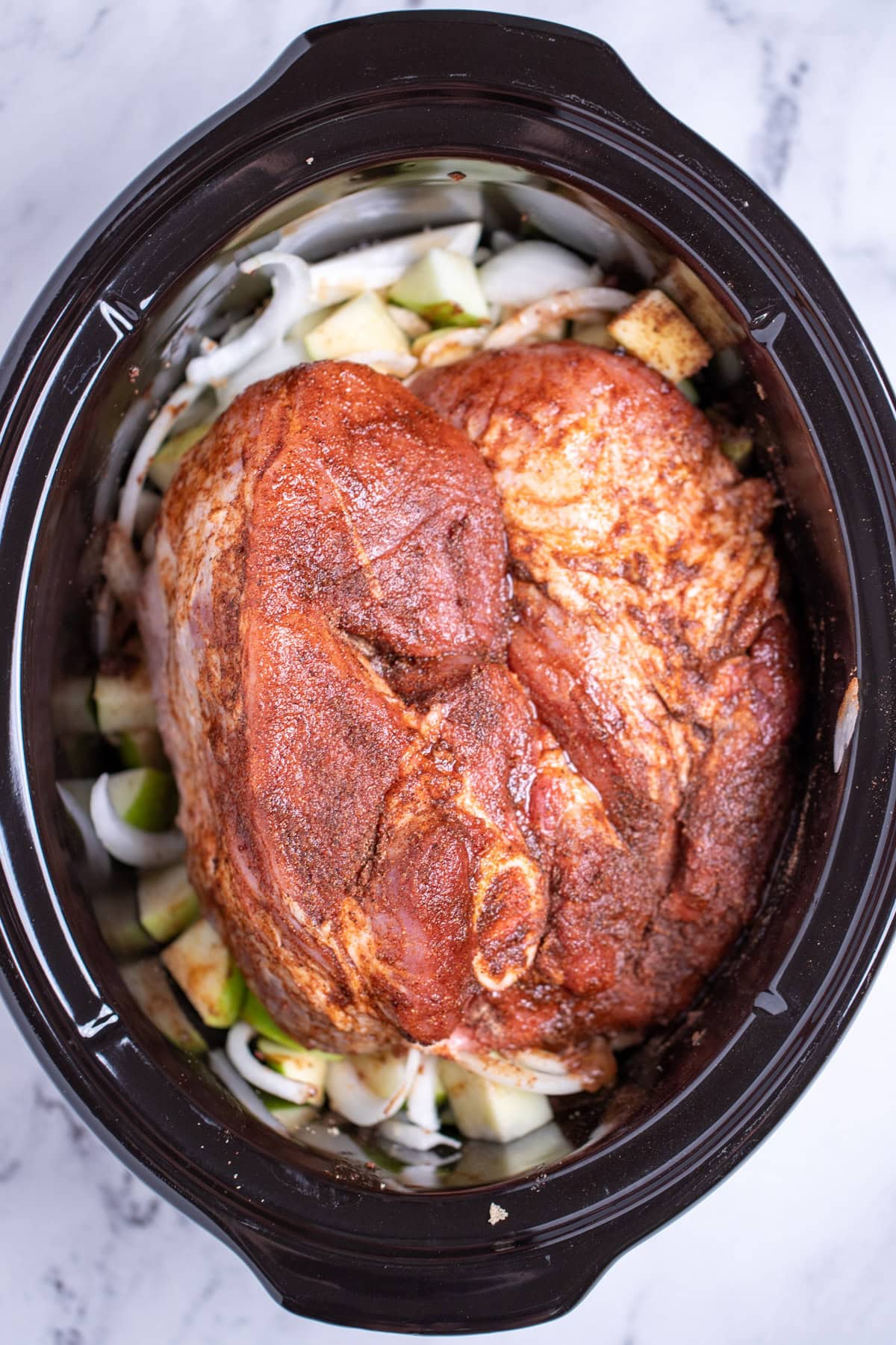 A raw pork shoulder rubbed in spices and oil in a slow cooker on top of apples and onions before being cooked.