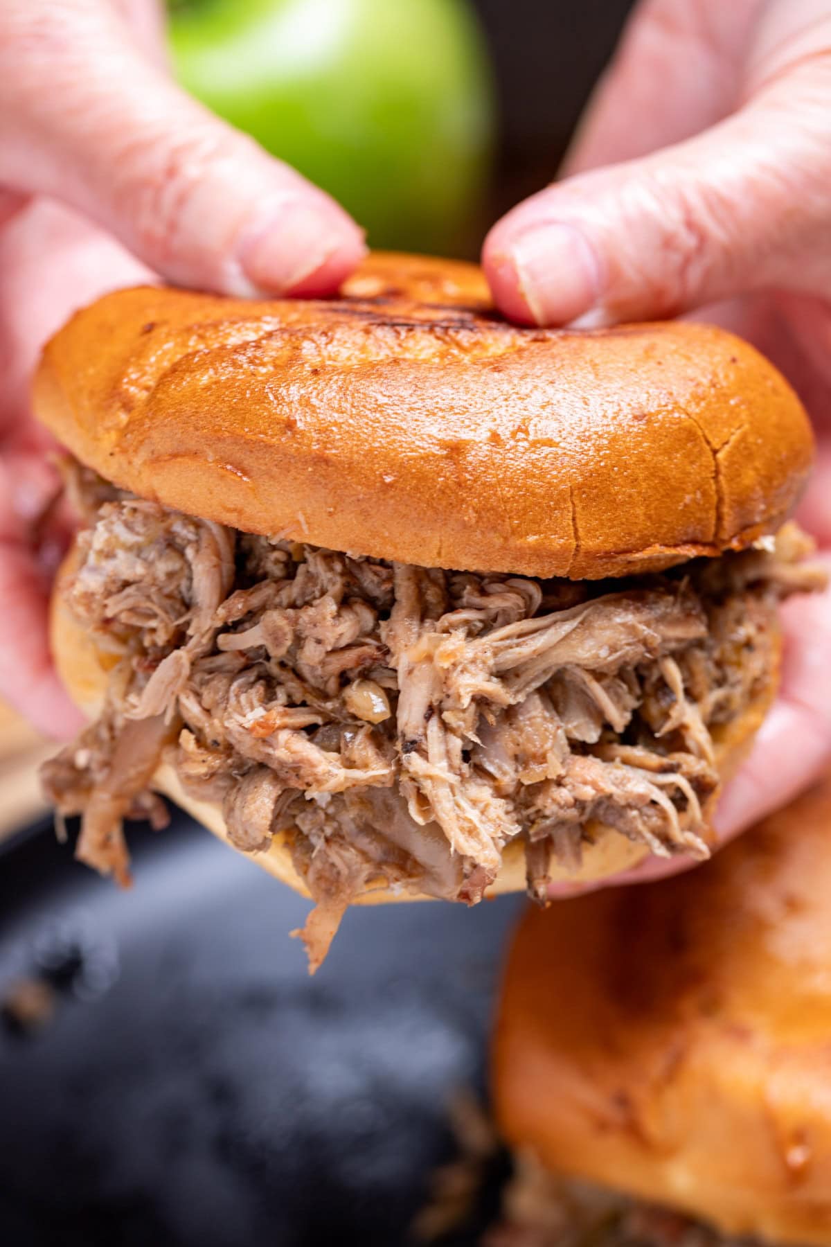 Two hands holding an apple cider pulled pork sandwich over a plate.