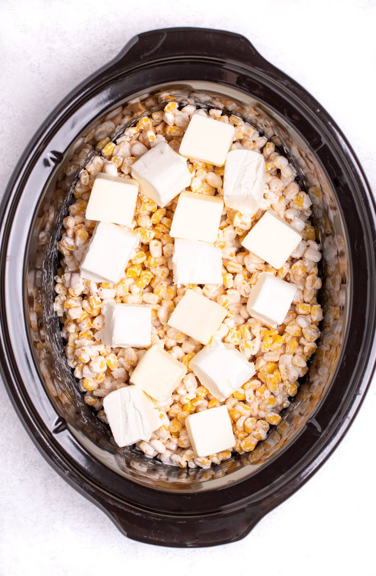 A slow cooker with frozen corn mixed with heavy cream, with pieces of butter and cream cheese on top, before being cooked.