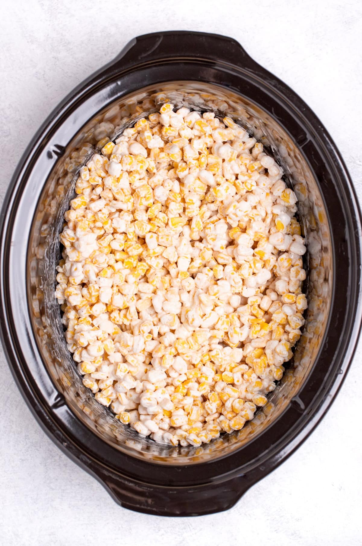A slow cooker with frozen corn kernels mixed with heavy cream and seasoning before being cooked.