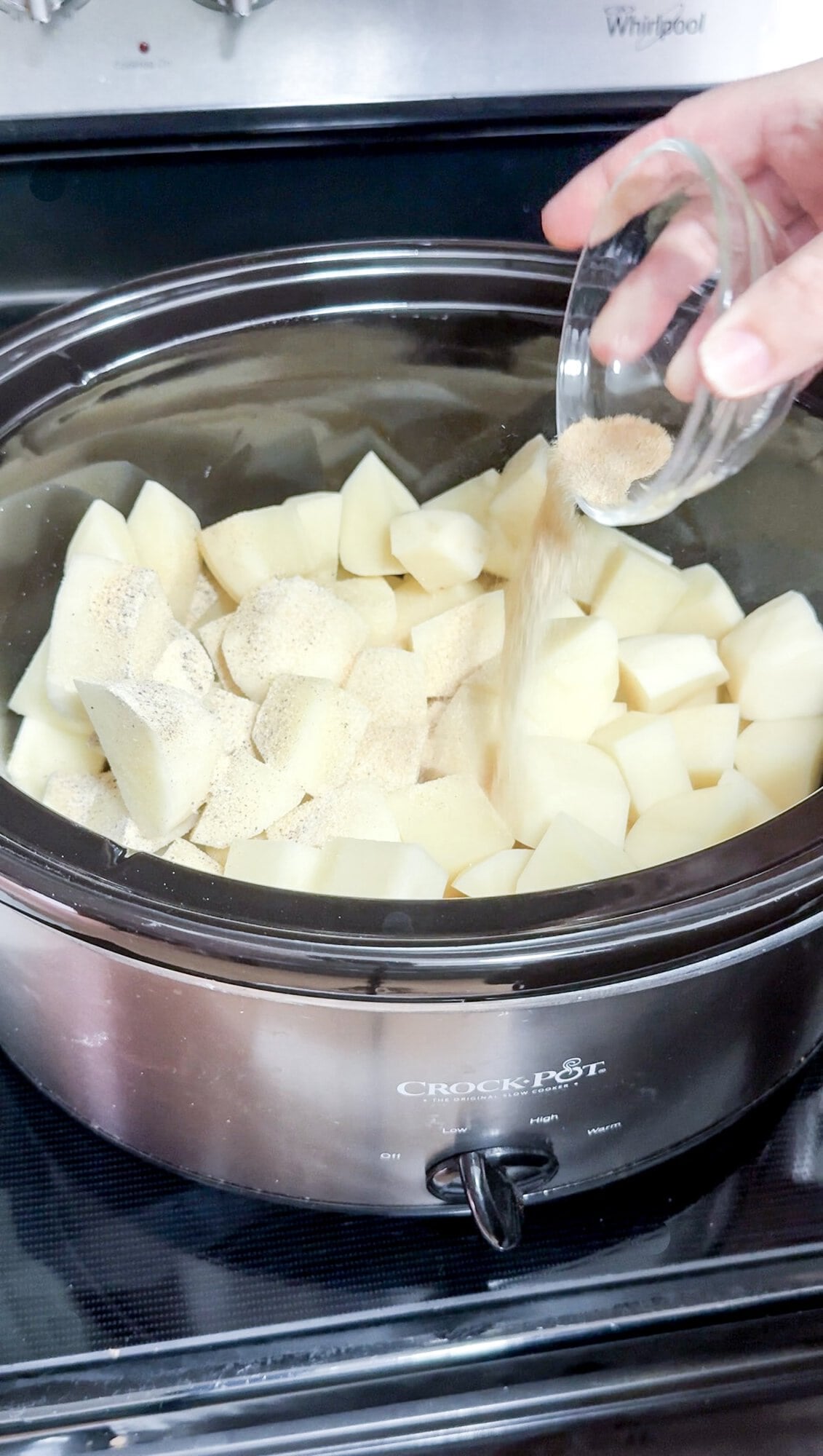 A slow cooker full of raw potato pieces, with a hand pouring spices over them.