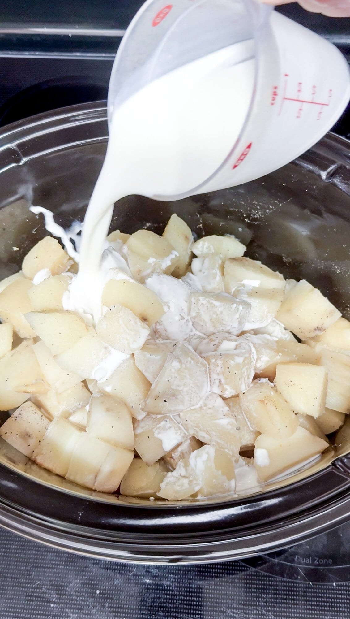 A slow cooker with cooked pieces of potatoes, and a hand pouring cream over them before mashing.