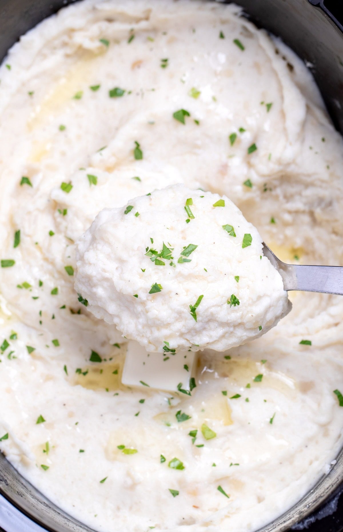 A slow cooker full of mashed potatoes topped with chopped parsley and butter, and a spoon lifting some up.