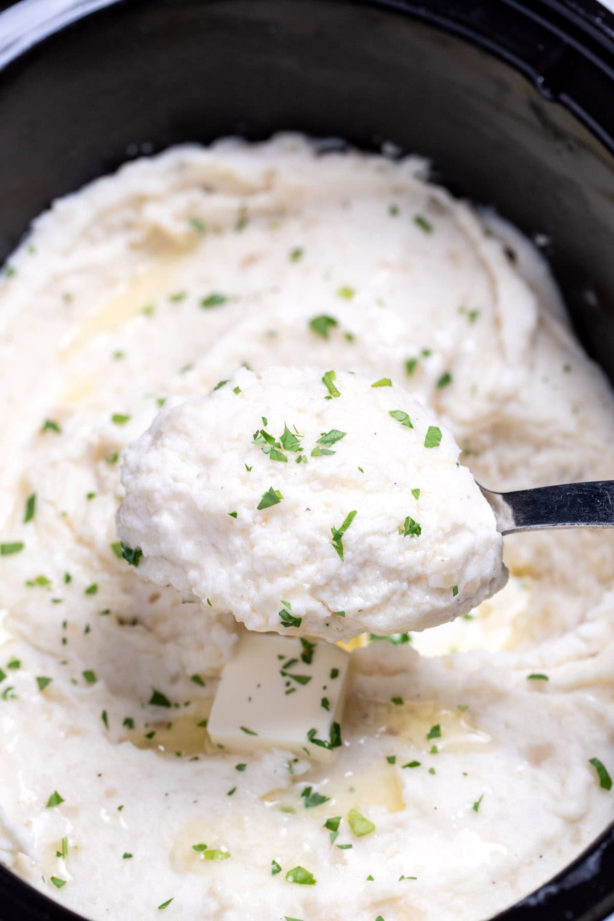 Mashed potatoes in a slow cooker topped with fresh parsley, and a spoon lifting some up.