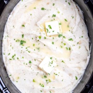 A slow cooker full of mashed potatoes topped with chopped parsley and butter.