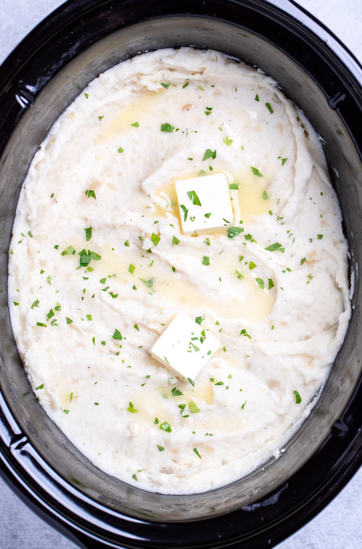A slow cooker full of mashed potatoes topped with chopped parsley and butter.