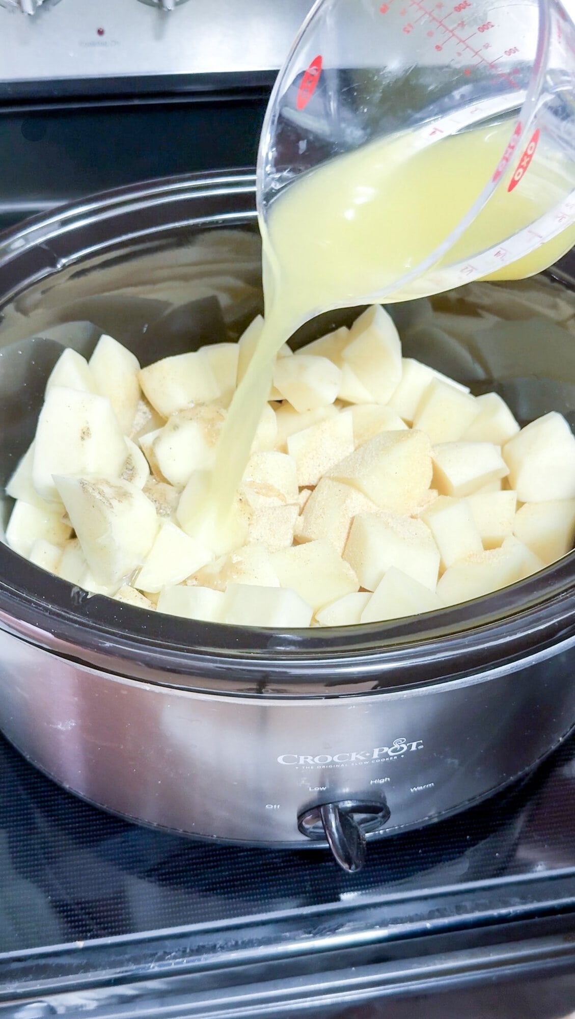 A slow cooker full of raw potato pieces, with a hand pouring broth over them, before cooking.
