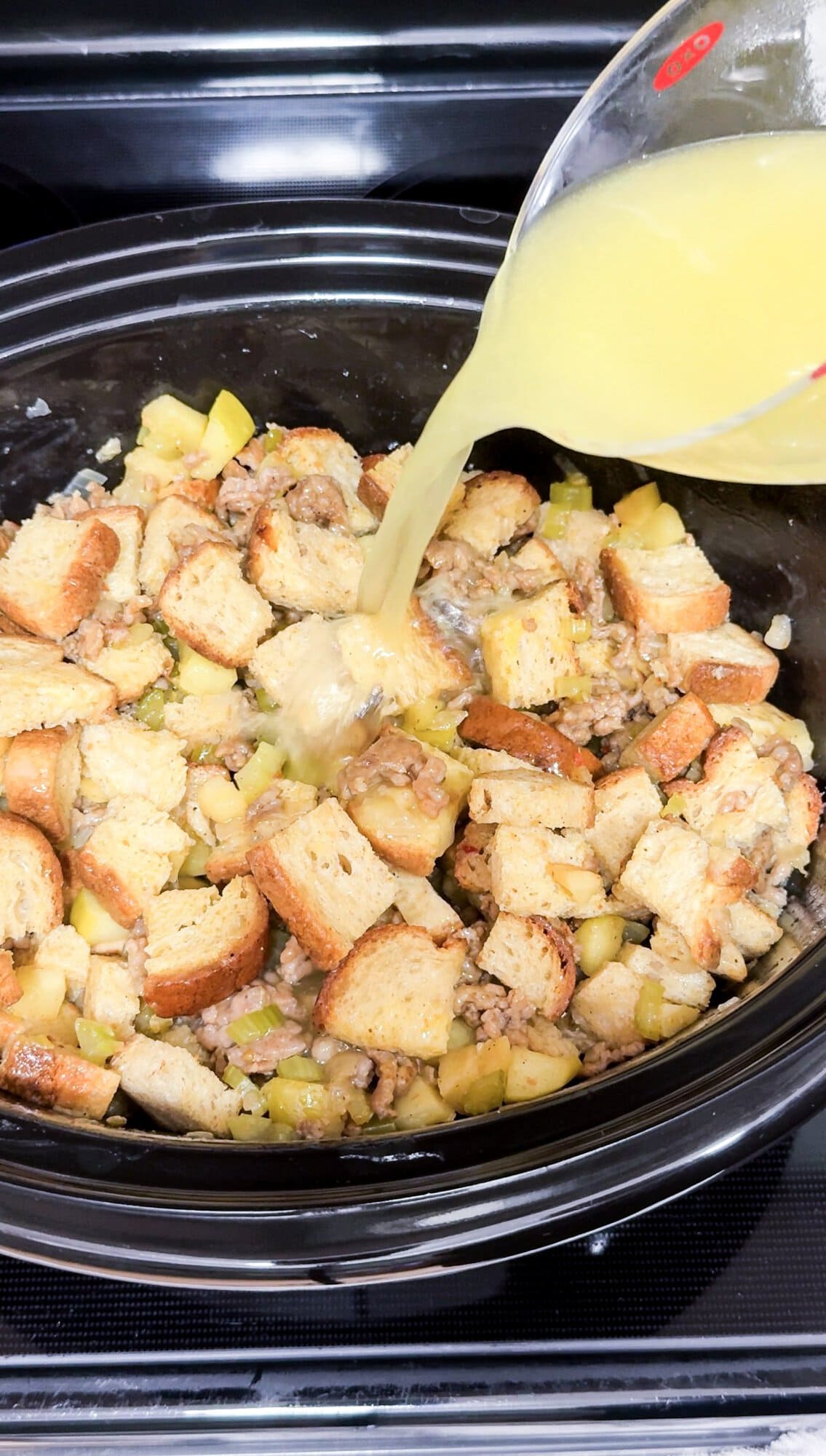 A slow cooker with gluten free sausage stuffing and broth being poured into it from a measuring cup, before cooking.