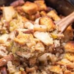 A slow cooker with a wooden spoon lifting gluten free sausage stuffing up.
