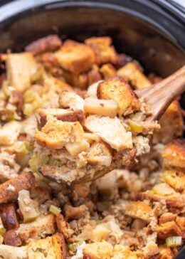 A slow cooker with a wooden spoon lifting gluten free sausage stuffing up.